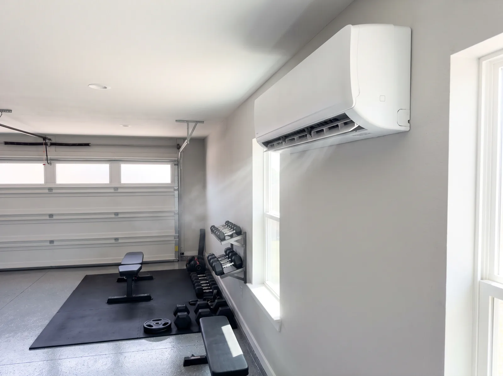 A ductless mini-split indoor head mounted high on the wall of a converted garage set up as a home gym, running above the equipment below
