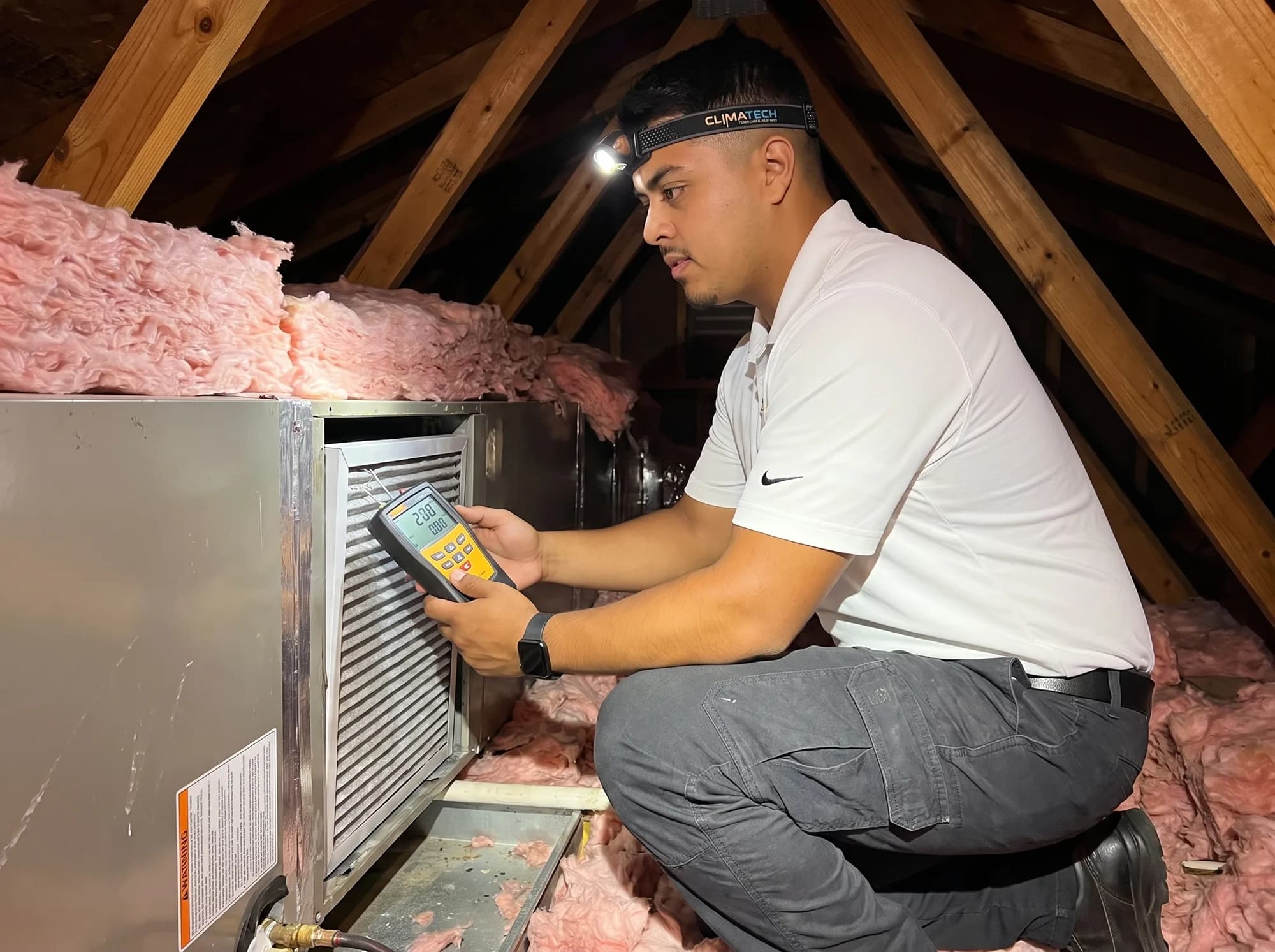 A technician kneeling in an attic, reading a handheld meter beside the return of an air handler